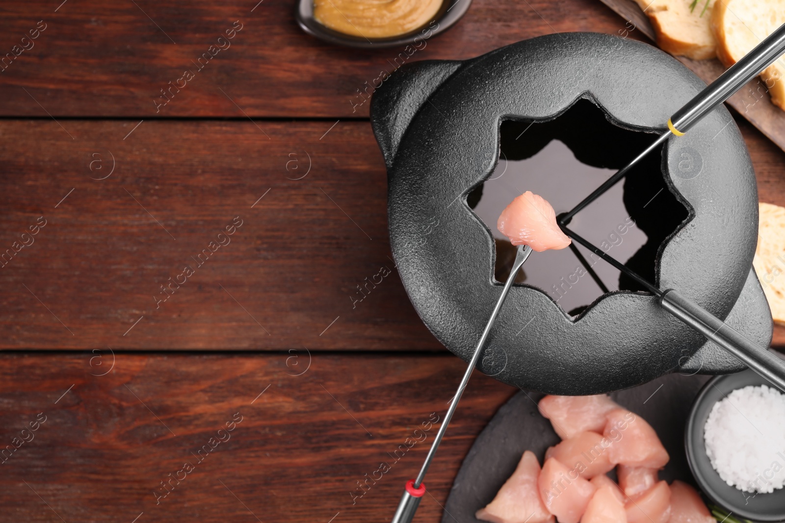 Photo of Fondue pot, forks, pieces of raw meat, bread and spices on wooden table, flat lay. Space for text