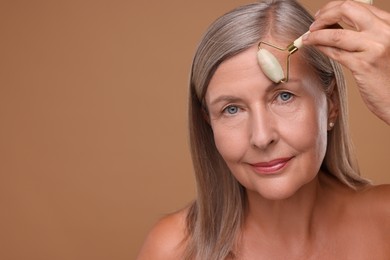 Photo of Woman massaging her face with jade roller on brown background. Space for text