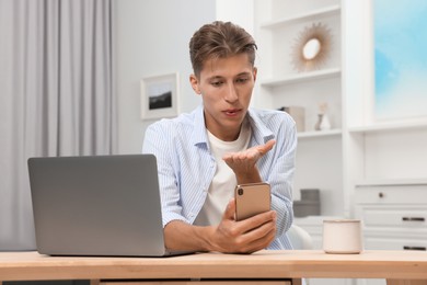 Happy young man having video chat via smartphone and blowing kiss at wooden table indoors. Long-distance relationship