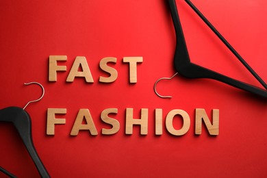 Phrase FAST FASHION made of wooden letters and black hangers on red background, flat lay