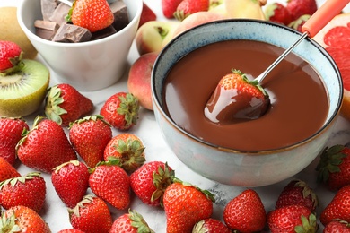 Photo of Fondue fork with strawberry in bowl of melted chocolate surrounded by other fruits on table