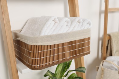 Photo of Soft towels in wicker basket on decorative ladder near white wall