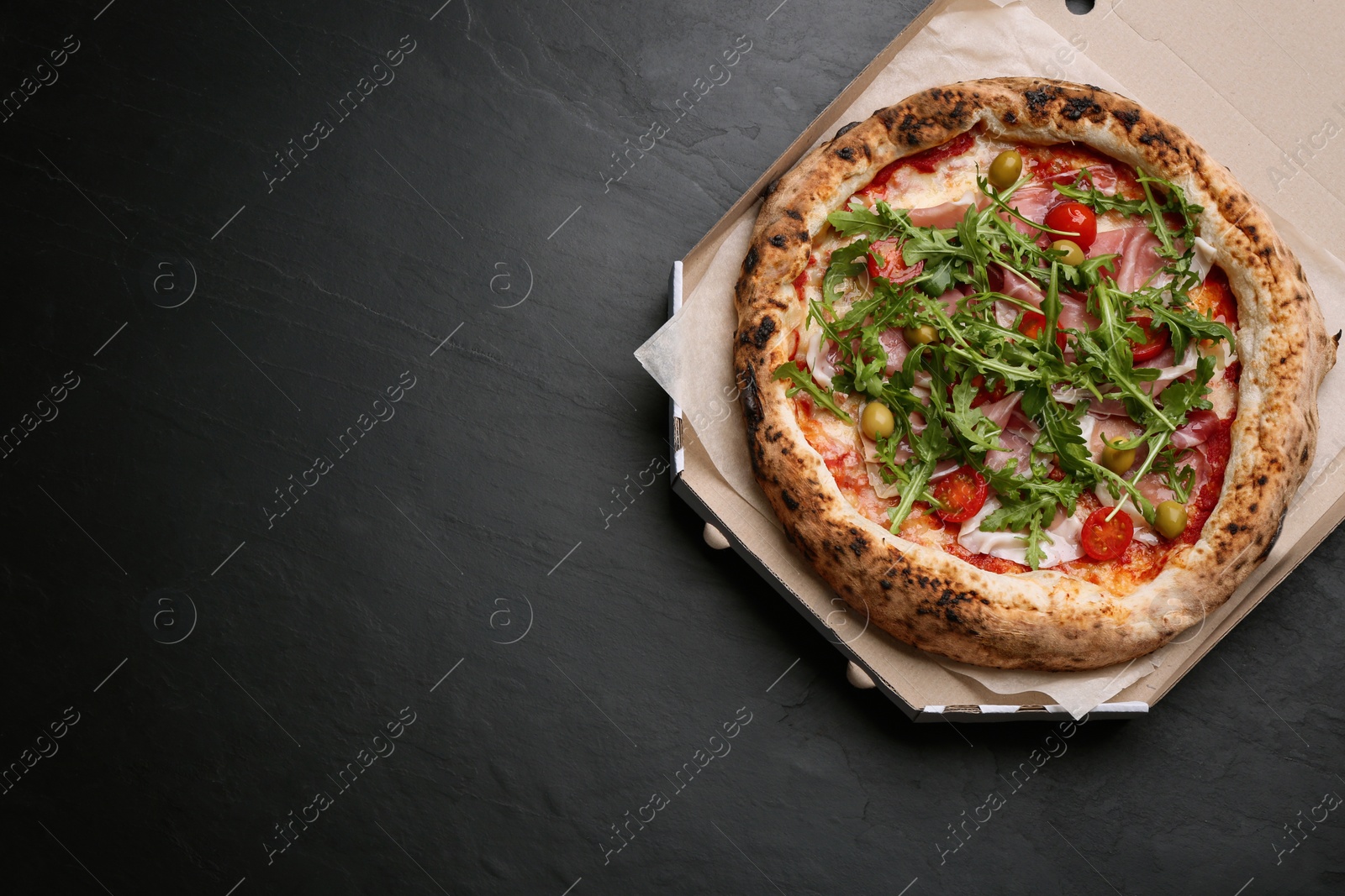 Photo of Tasty pizza with meat and arugula in cardboard box on black table, top view. Space for text