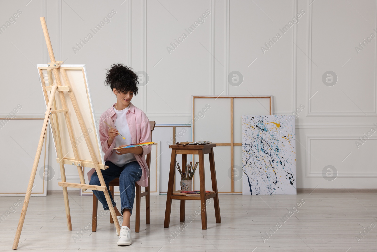 Photo of Young woman holding brush and artist`s palette near easel with canvas