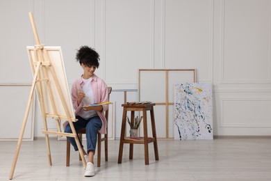 Photo of Young woman holding brush and artist`s palette near easel with canvas