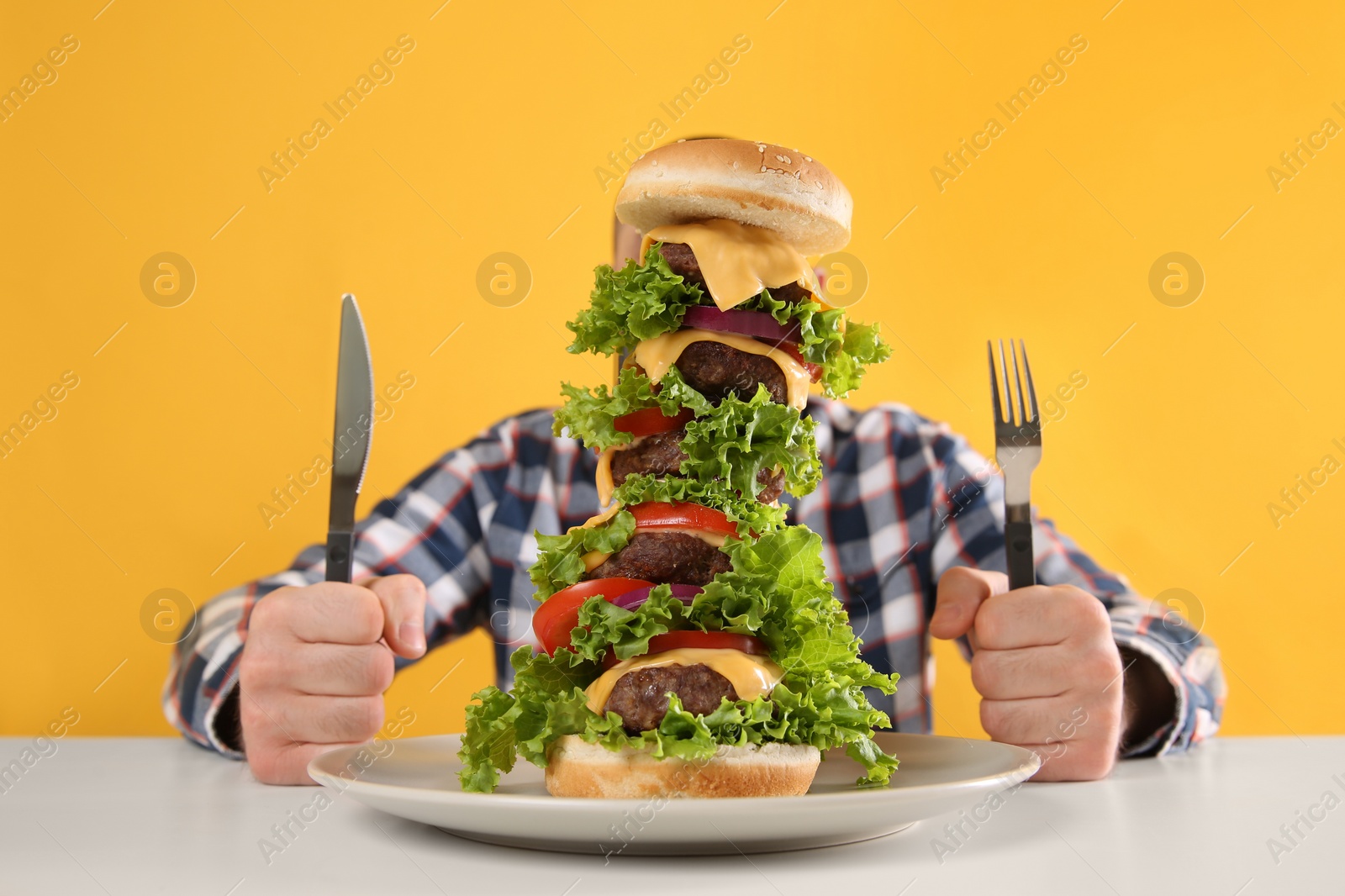Photo of Hungry man with cutlery and huge burger at white table on yellow background