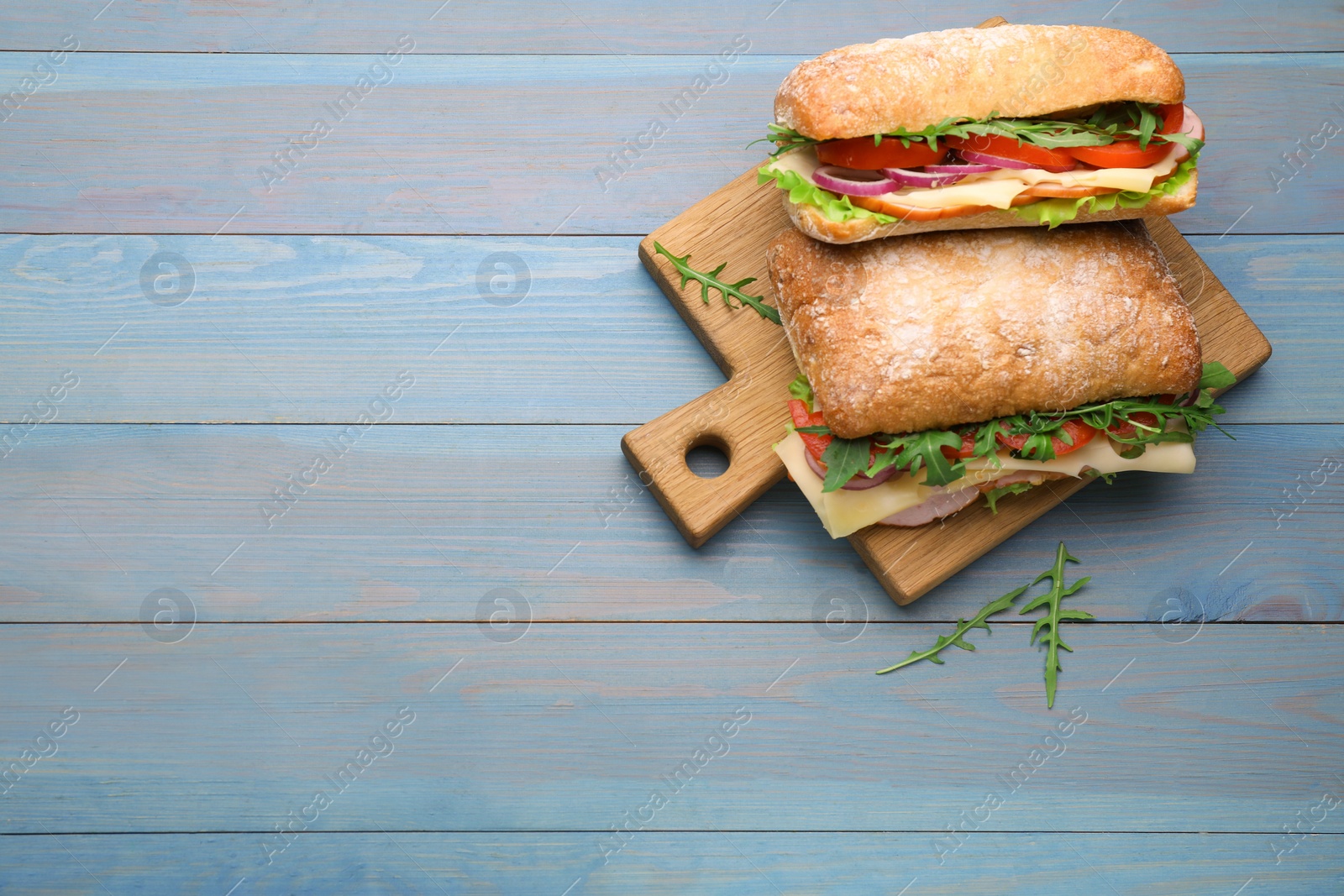 Photo of Delicious sandwiches with fresh vegetables, cheese and arugula on light blue wooden table, flat lay. Space for text