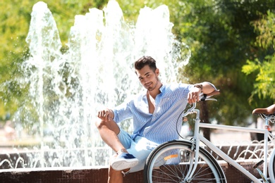 Photo of Handsome young hipster man sitting with bicycle near fountain in park