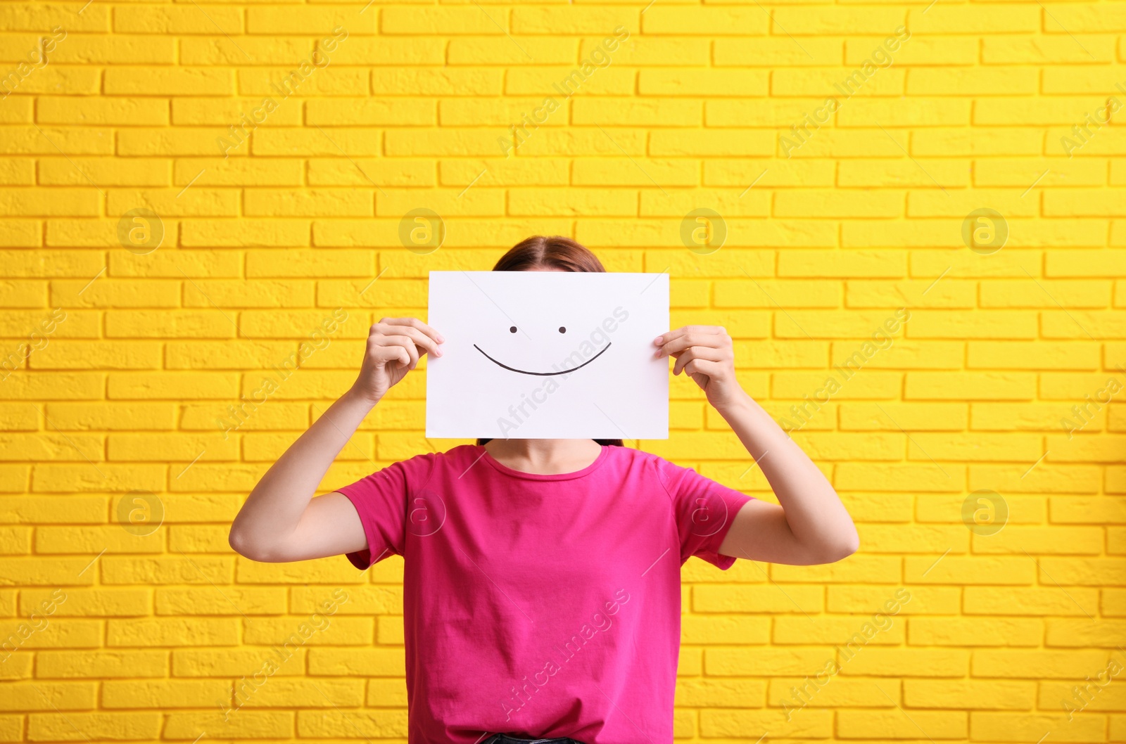 Photo of Woman hiding behind sheet of paper with happy face against yellow brick wall