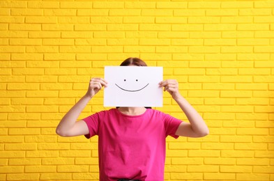 Photo of Woman hiding behind sheet of paper with happy face against yellow brick wall