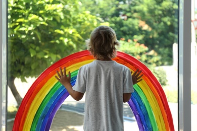 Little boy near rainbow painting on window indoors, back view. Stay at home concept