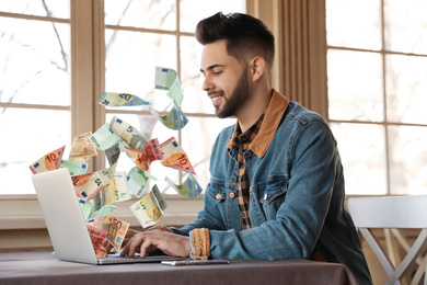 Image of Man with modern laptop and flying euro banknotes at table indoors. People make money online