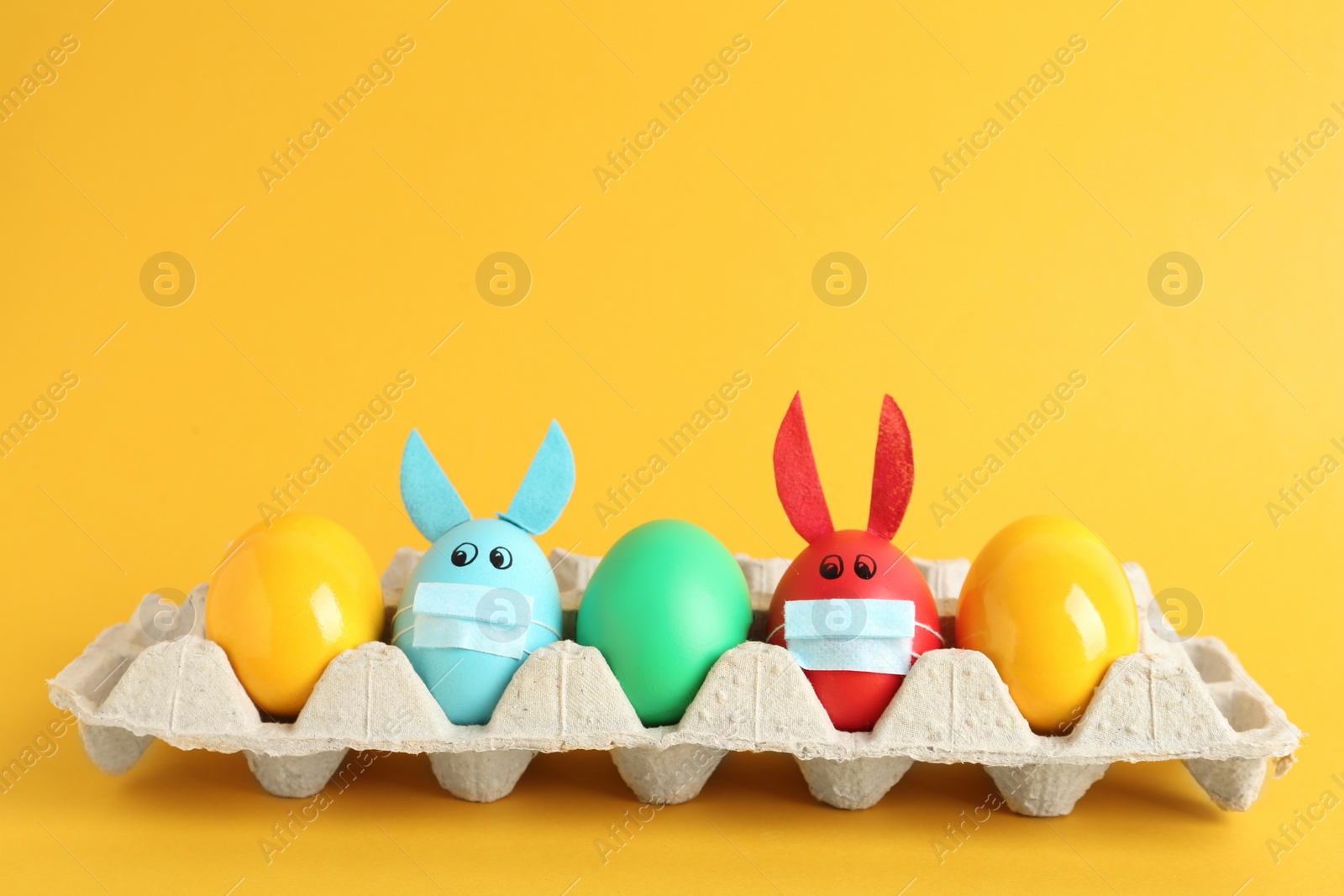 Photo of Painted eggs decorated with bunny ears and protective masks in package on yellow background. Easter holiday during COVID-19 quarantine