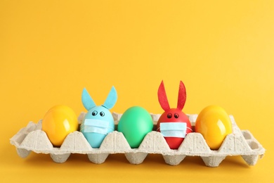 Photo of Painted eggs decorated with bunny ears and protective masks in package on yellow background. Easter holiday during COVID-19 quarantine