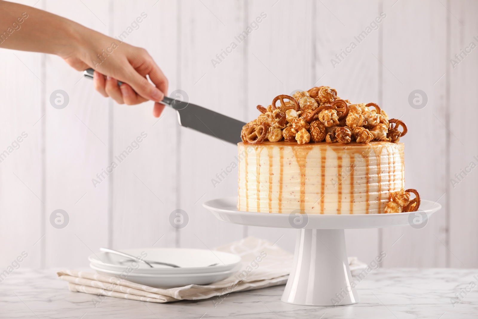 Photo of Woman cutting caramel drip cake decorated with popcorn and pretzels at white marble table, closeup