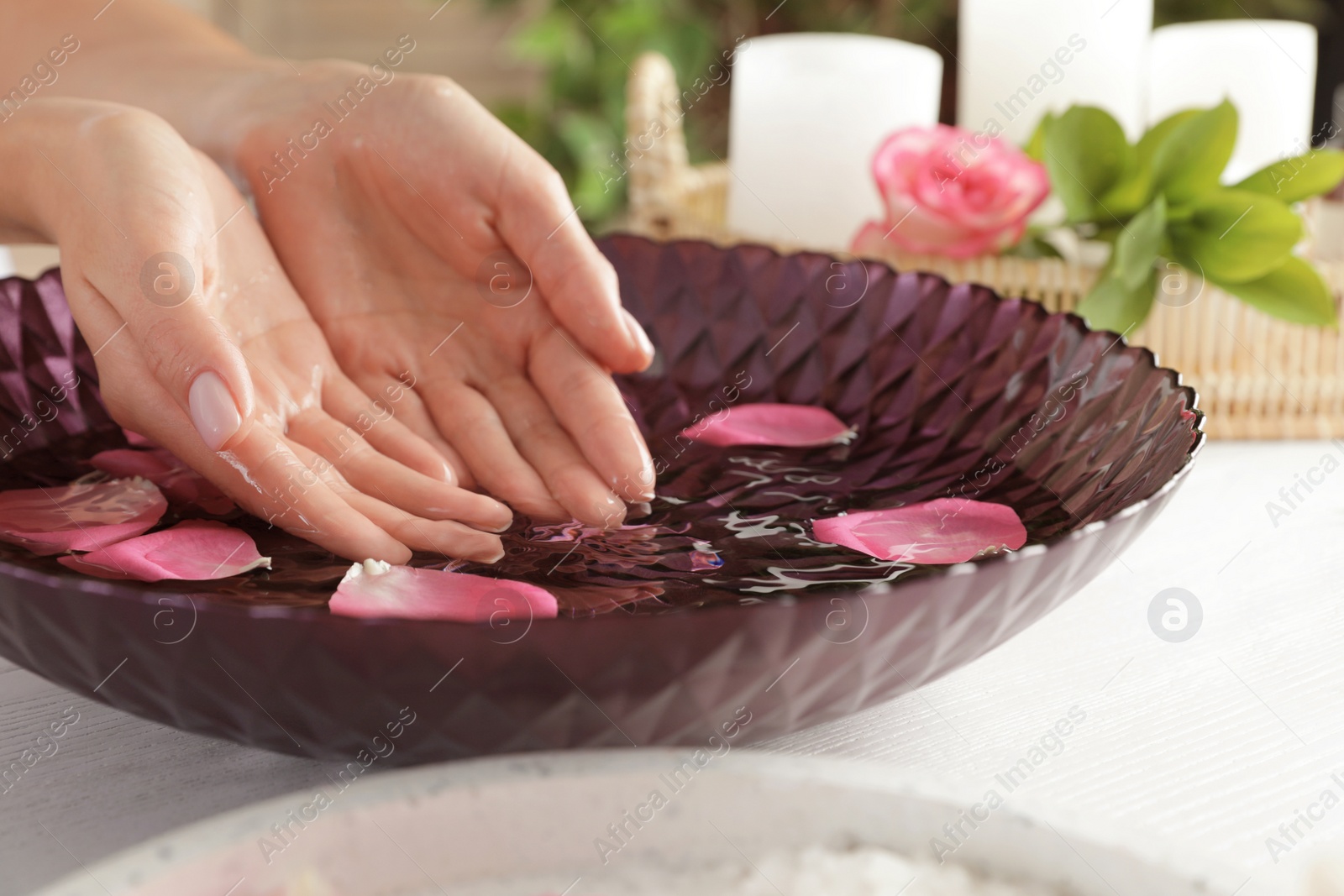 Photo of Woman soaking her hands in bowl of water and petals on table, closeup with space for text. Spa treatment