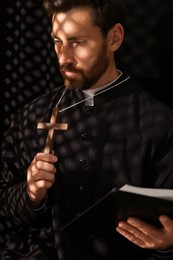 Catholic priest in cassock holding cross with Bible in confessional booth