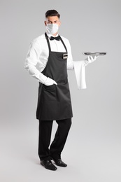 Waiter in medical face mask with empty tray on light grey background
