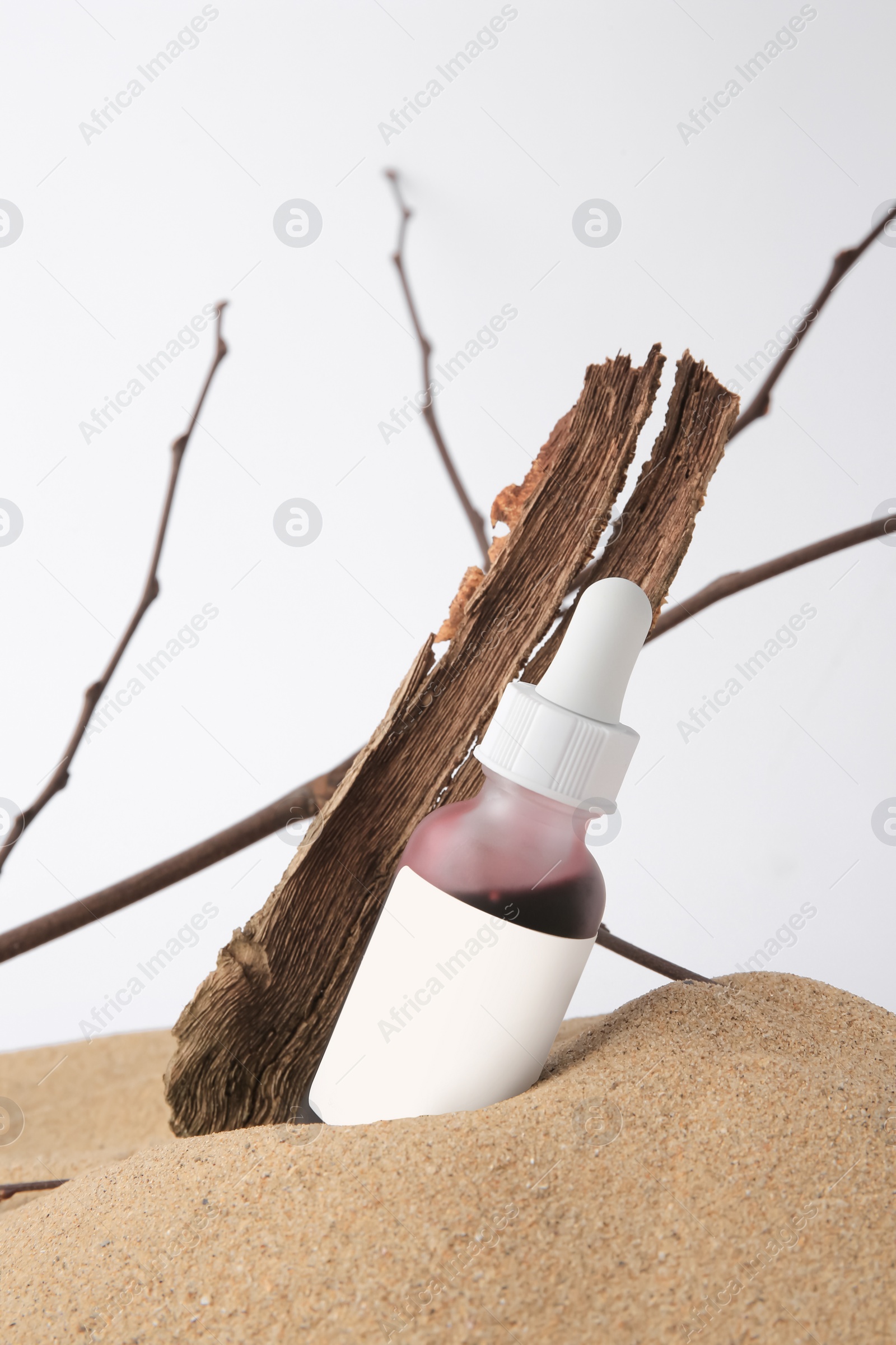 Photo of Bottle with serum, bark and branches on sand against white background. Cosmetic product