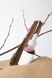 Bottle with serum, bark and branches on sand against white background. Cosmetic product
