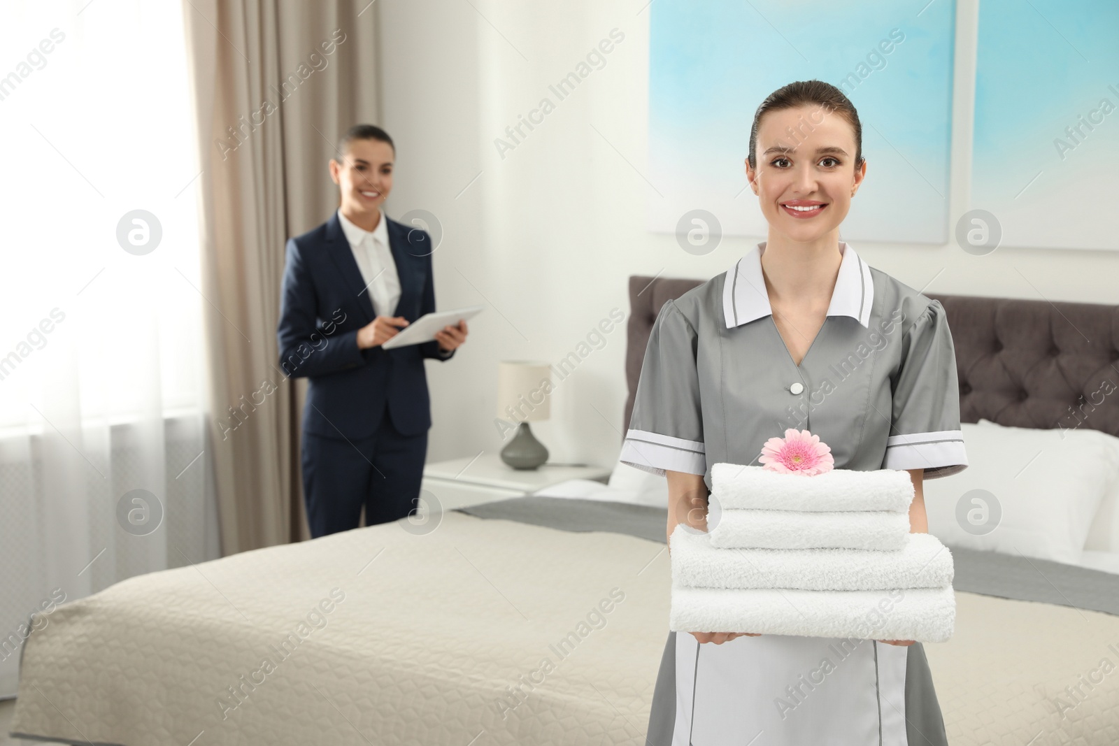Photo of Chambermaid with stack of fresh towels and blurred supervisor in hotel room. Space for text