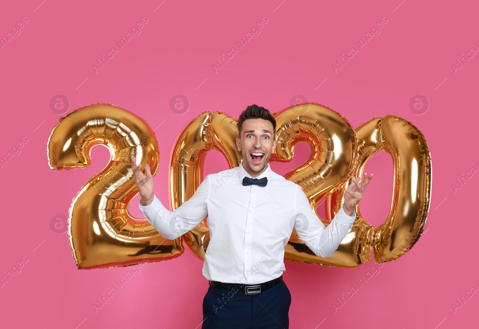 Photo of Happy young man near golden 2020 balloons on pink background. New Year celebration