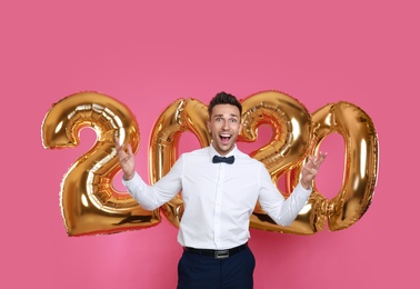 Photo of Happy young man near golden 2020 balloons on pink background. New Year celebration