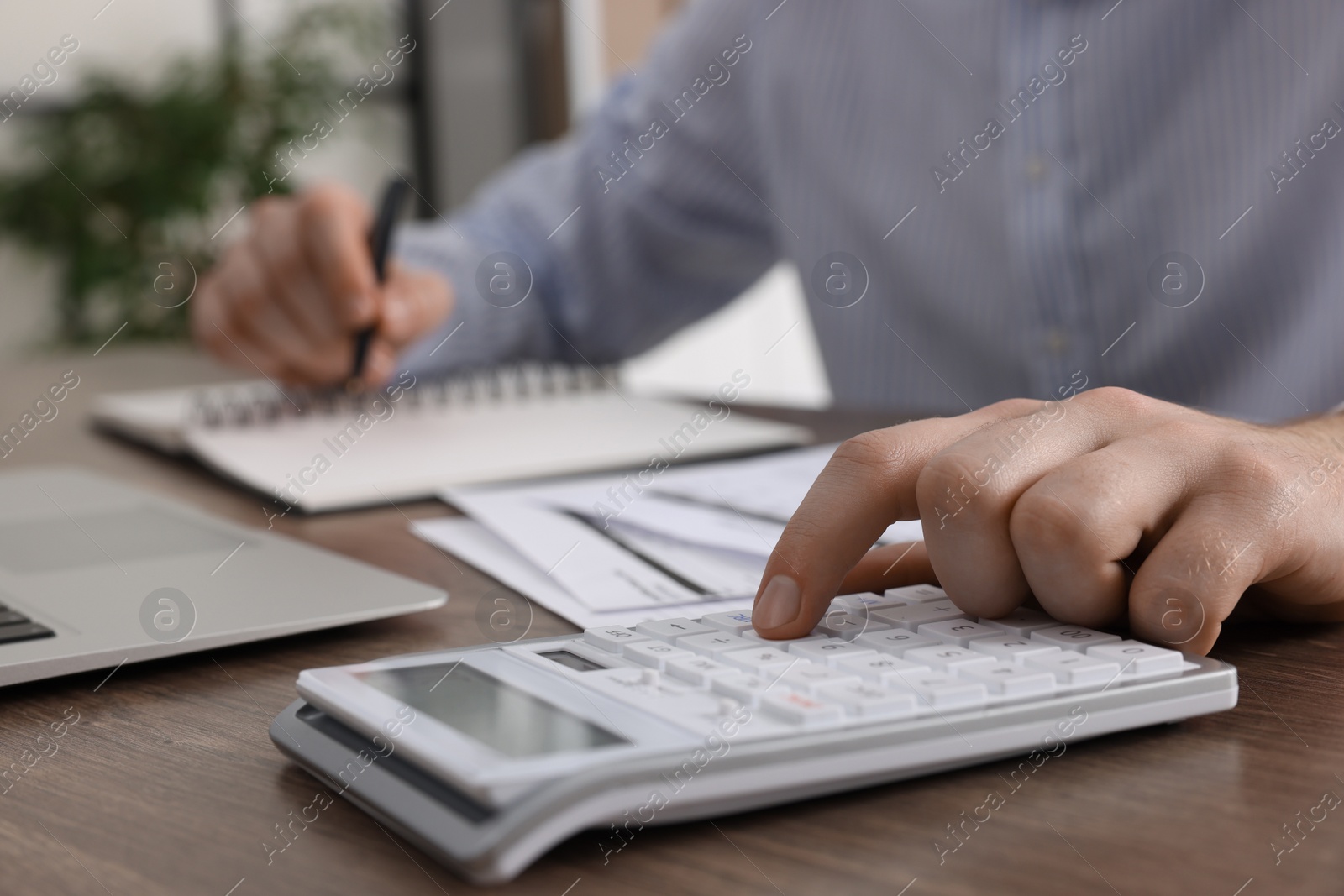 Photo of Man using calculator while taking notes at wooden table indoors, closeup