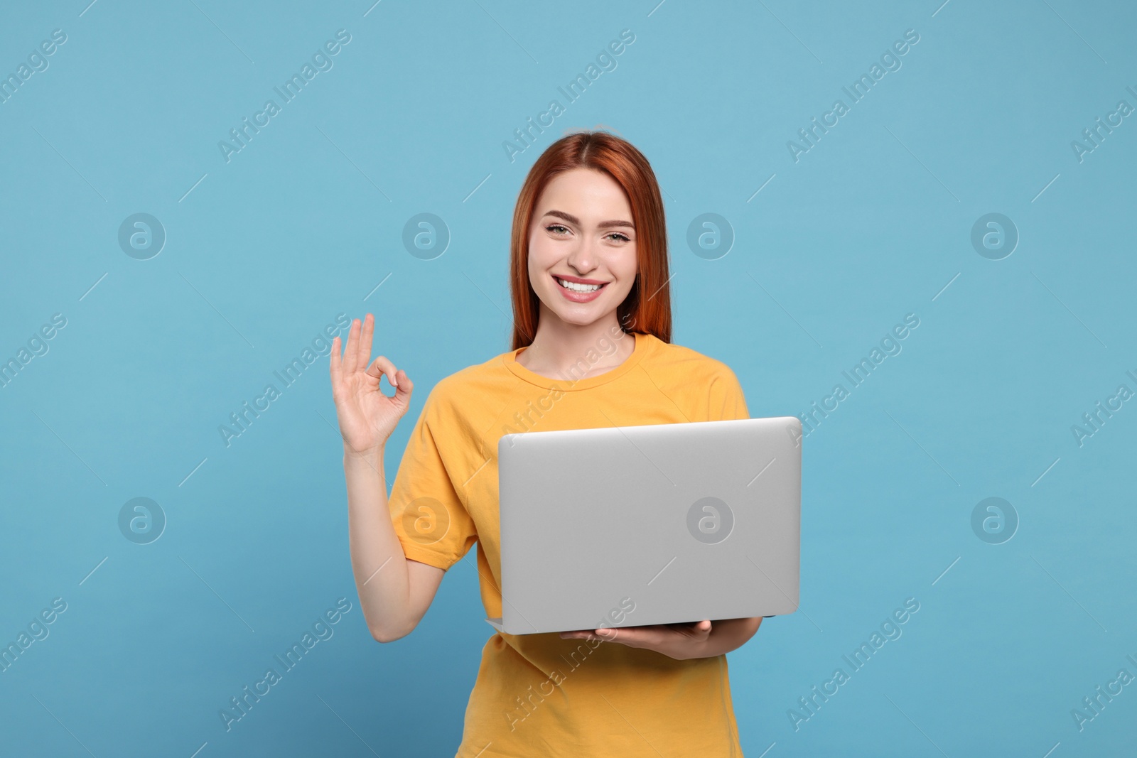 Photo of Smiling young woman with laptop showing OK gesture on light blue background