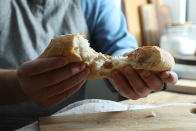 Photo of Man breaking loaf of fresh bread at wooden table indoors, closeup