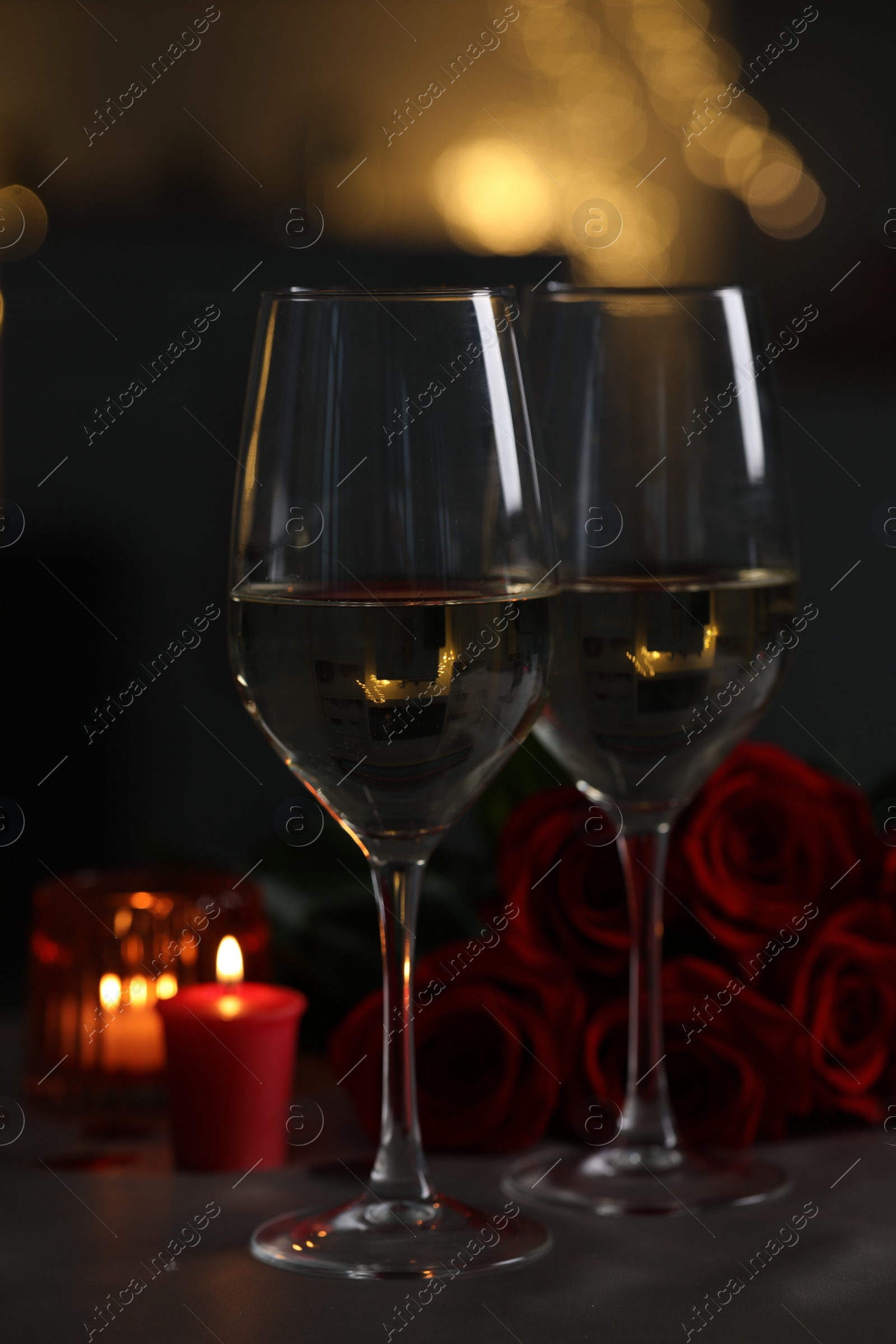 Photo of Glasses of white wine, burning candle and rose flowers on grey table against blurred lights. Romantic atmosphere