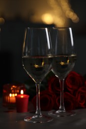 Glasses of white wine, burning candle and rose flowers on grey table against blurred lights. Romantic atmosphere