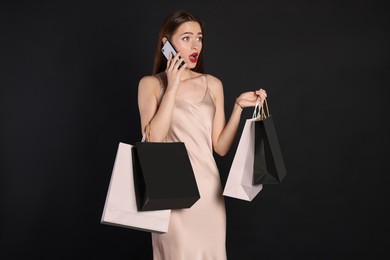 Photo of Surprised young woman with shopping bags and smartphone on black background. Big sale