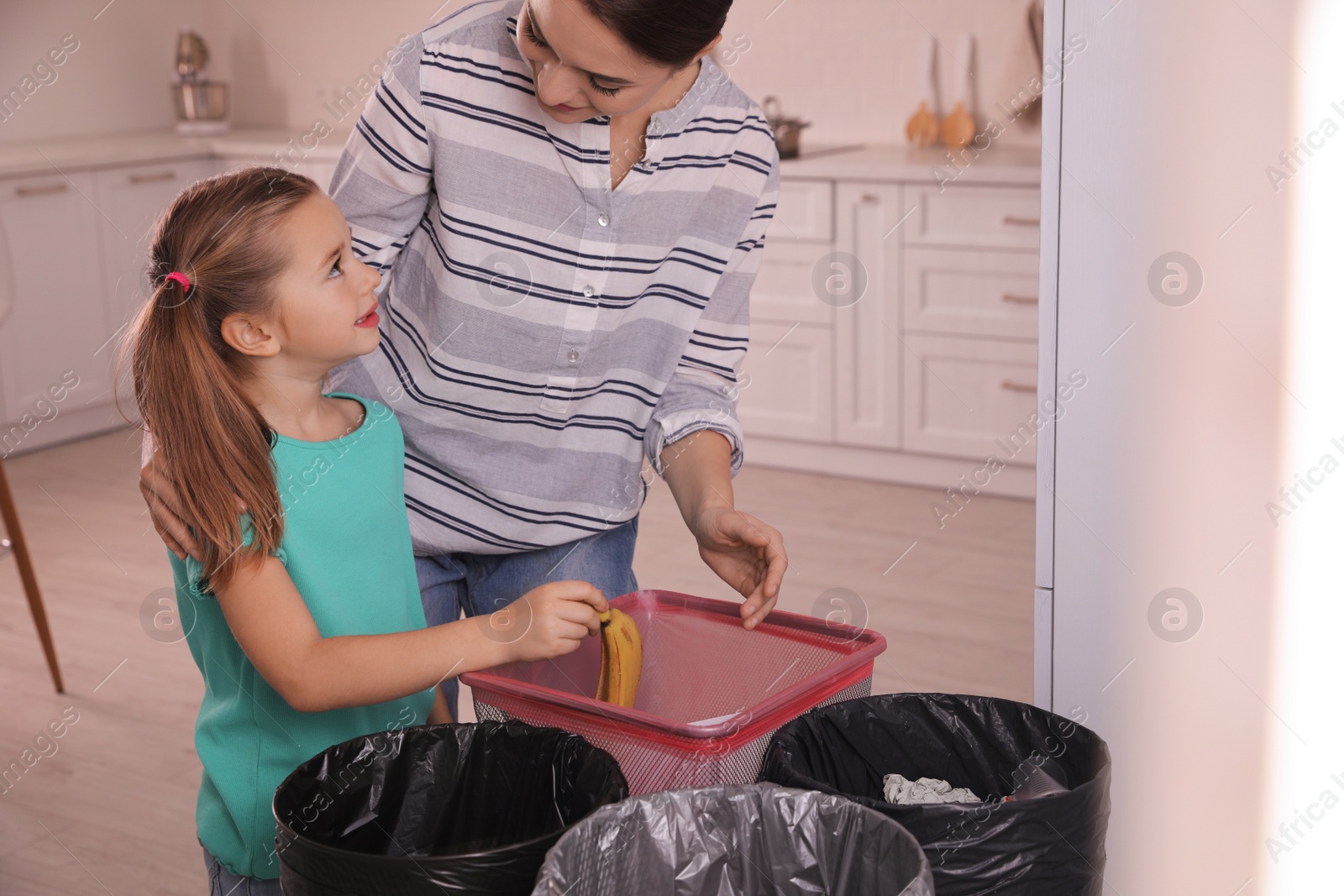 Photo of Young woman and her daughter throwing banana peel into trash bin in kitchen. Separate waste collection