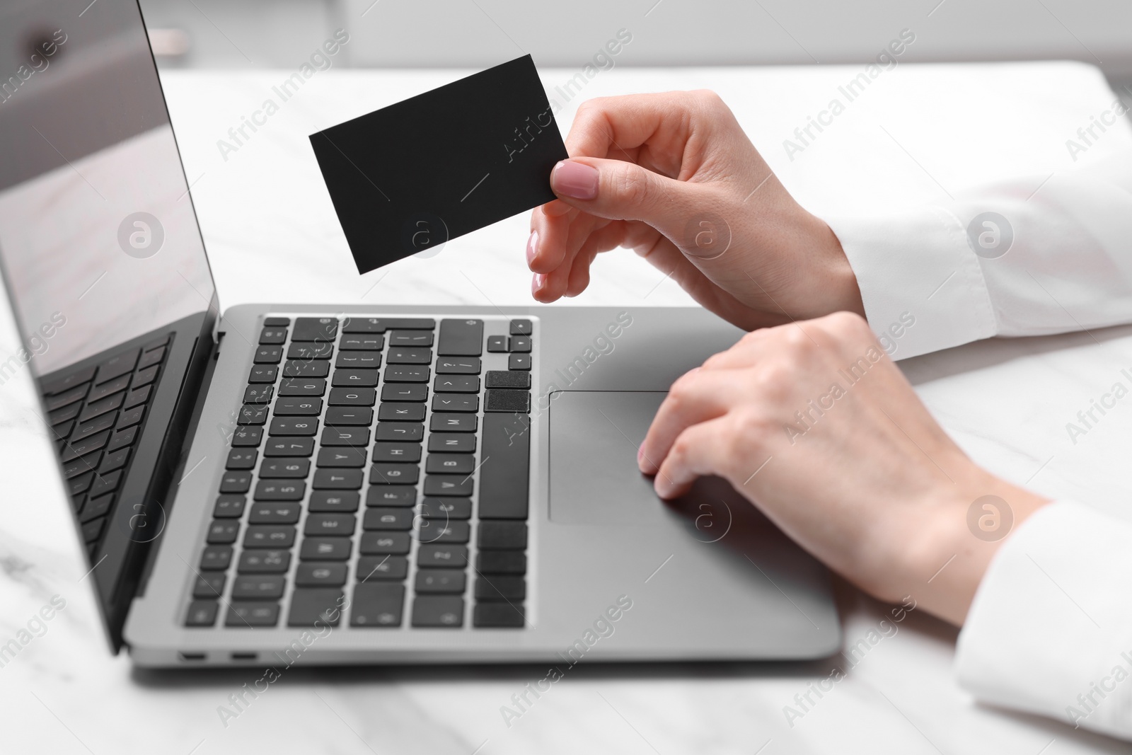 Photo of Woman with laptop holding blank business card at white table, closeup. Space for text