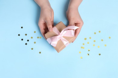 Photo of Woman holding gift box with pink bow and confetti on light blue background, top view