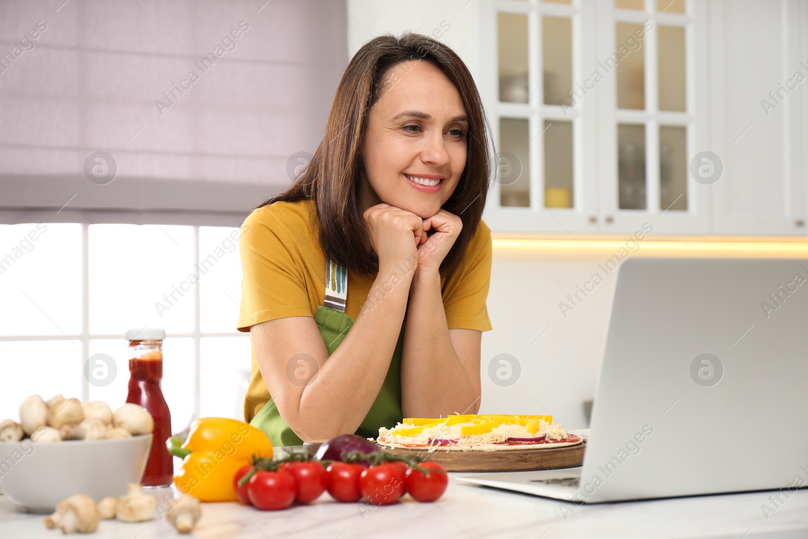 Photo of Happy woman making pizza while watching online cooking course via laptop in kitchen