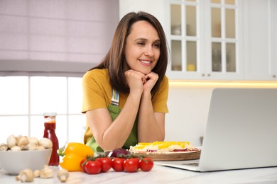 Happy woman making pizza while watching online cooking course via laptop in kitchen