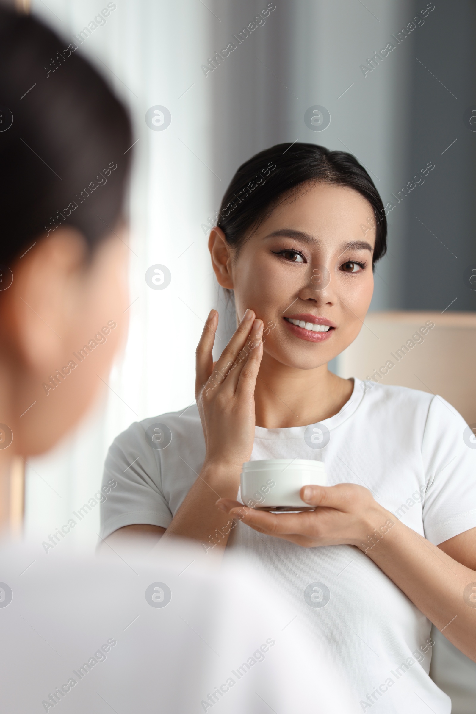 Photo of Happy woman applying face cream near mirror at home