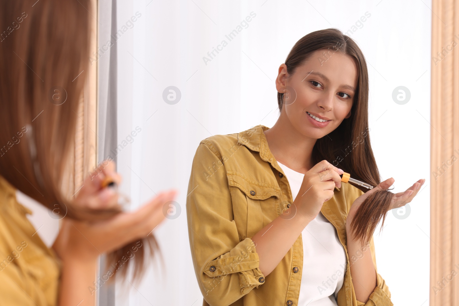 Photo of Beautiful woman applying serum onto hair near mirror indoors