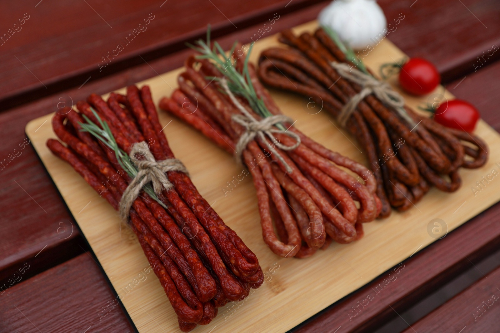 Photo of Bundles of delicious kabanosy with rosemary, tomatoes and garlic on wooden table