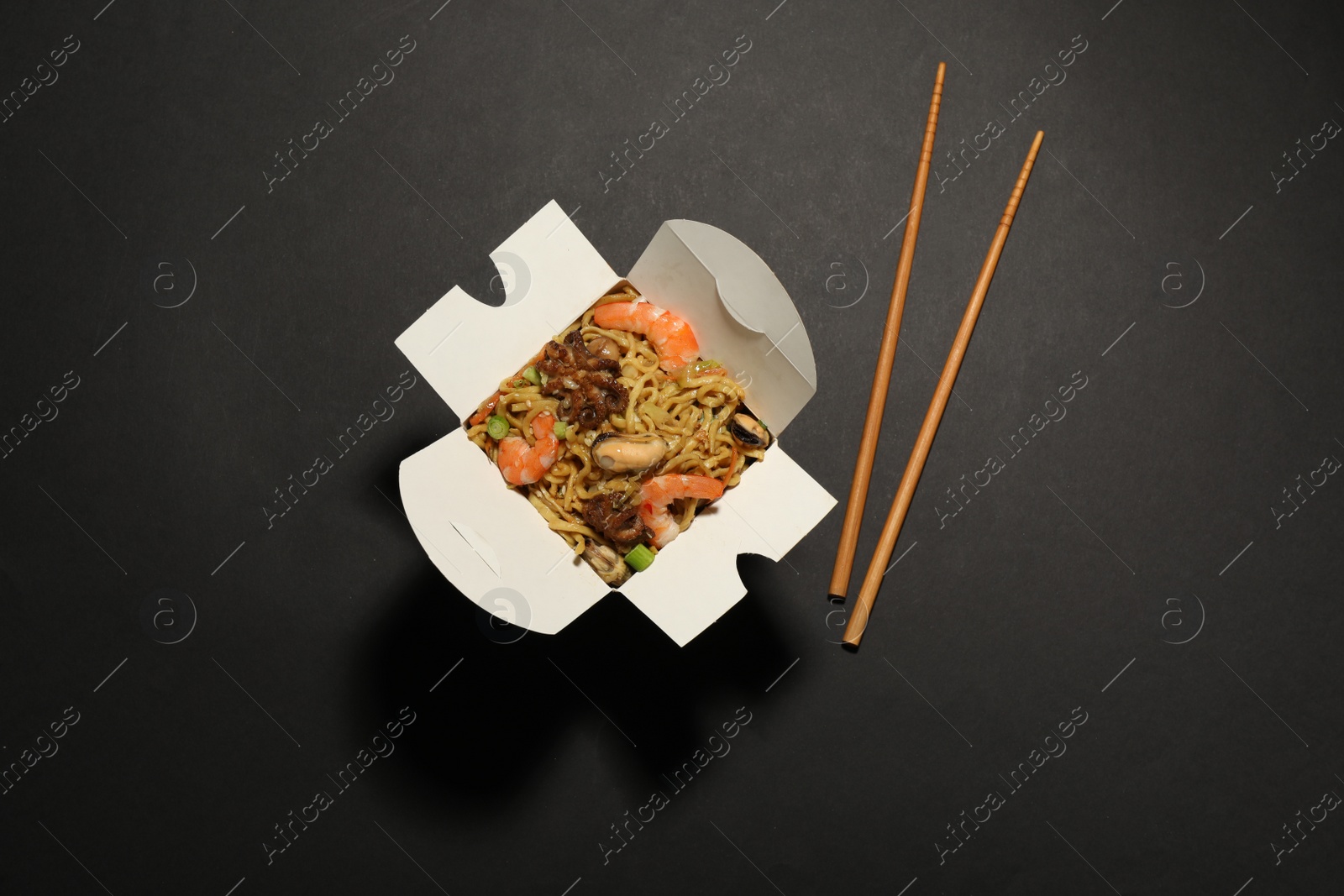 Photo of Box of wok noodles with seafood and chopsticks on black background, flat lay