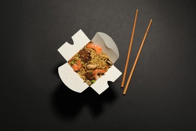 Photo of Box of wok noodles with seafood and chopsticks on black background, flat lay