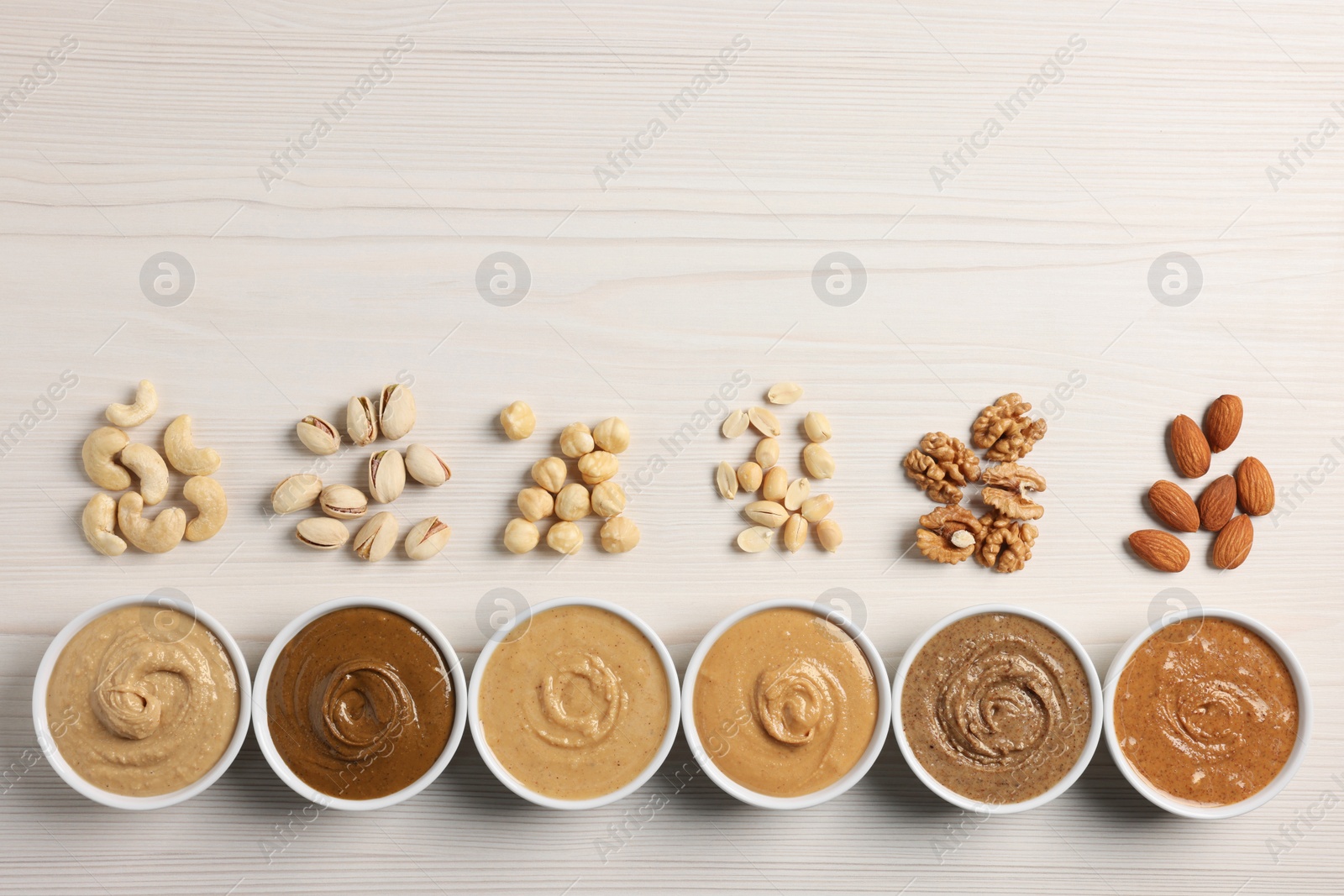 Photo of Many tasty nut butters in bowls and nuts on white wooden table, flat lay. Space for text
