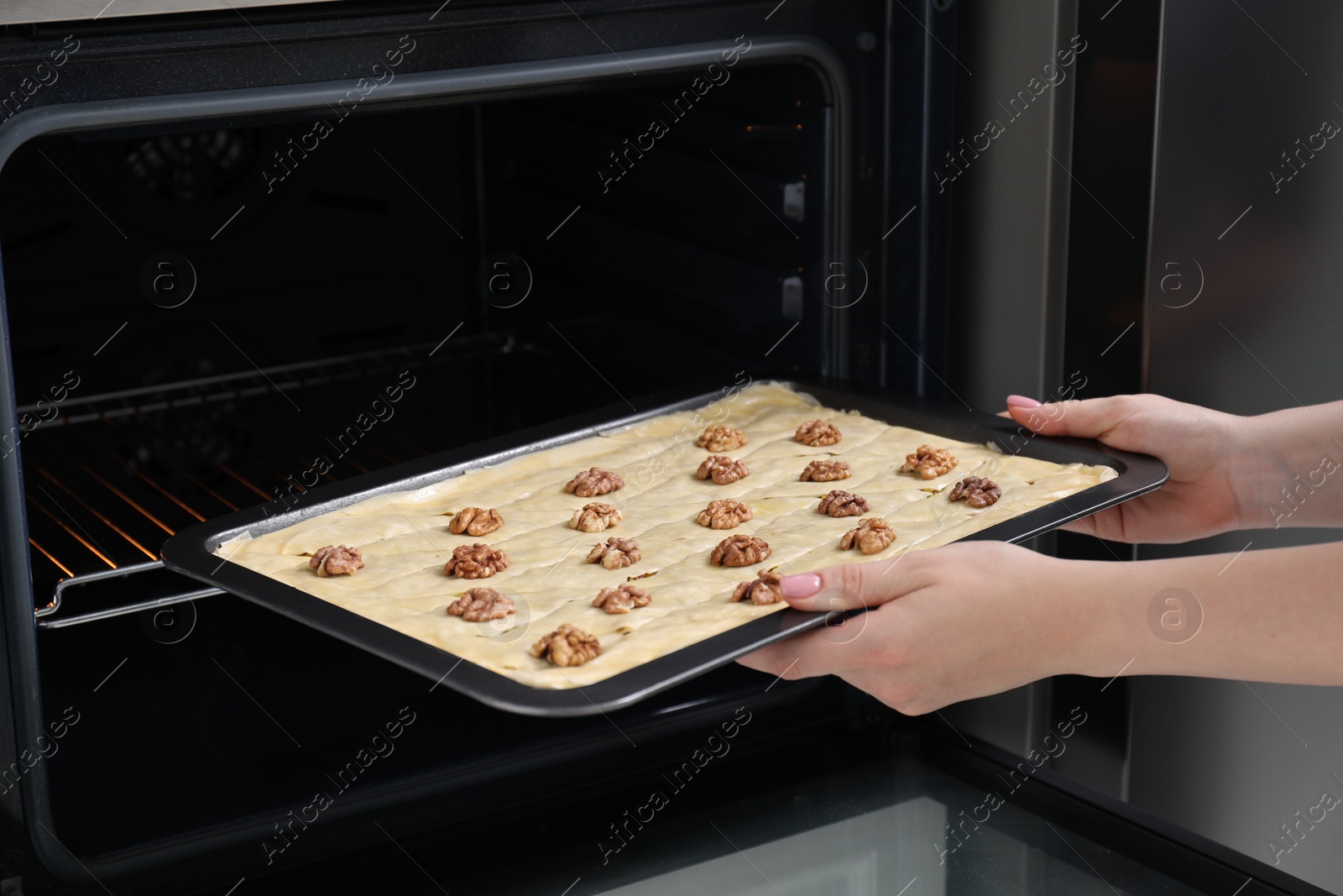 Photo of Woman putting baking pan with baklava into oven, closeup