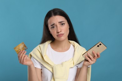 Confused woman with credit card and smartphone on light blue background. Debt problem