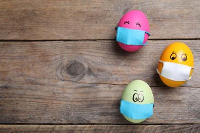 Bright eggs in protective masks on wooden table, flat lay with space for text. Easter holiday during coronavirus quarantine