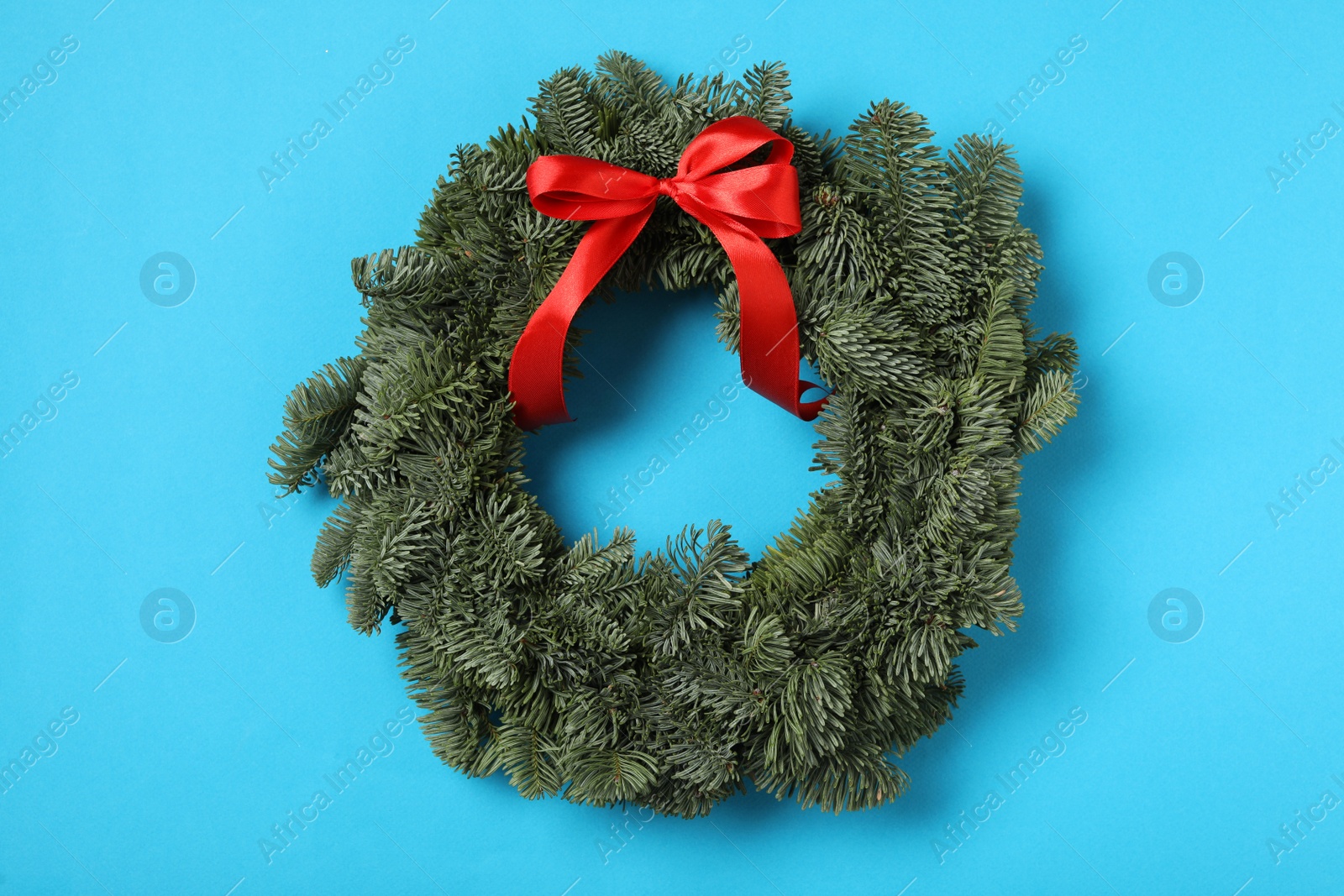 Photo of Christmas wreath made of fir tree branches with red ribbon on light blue background