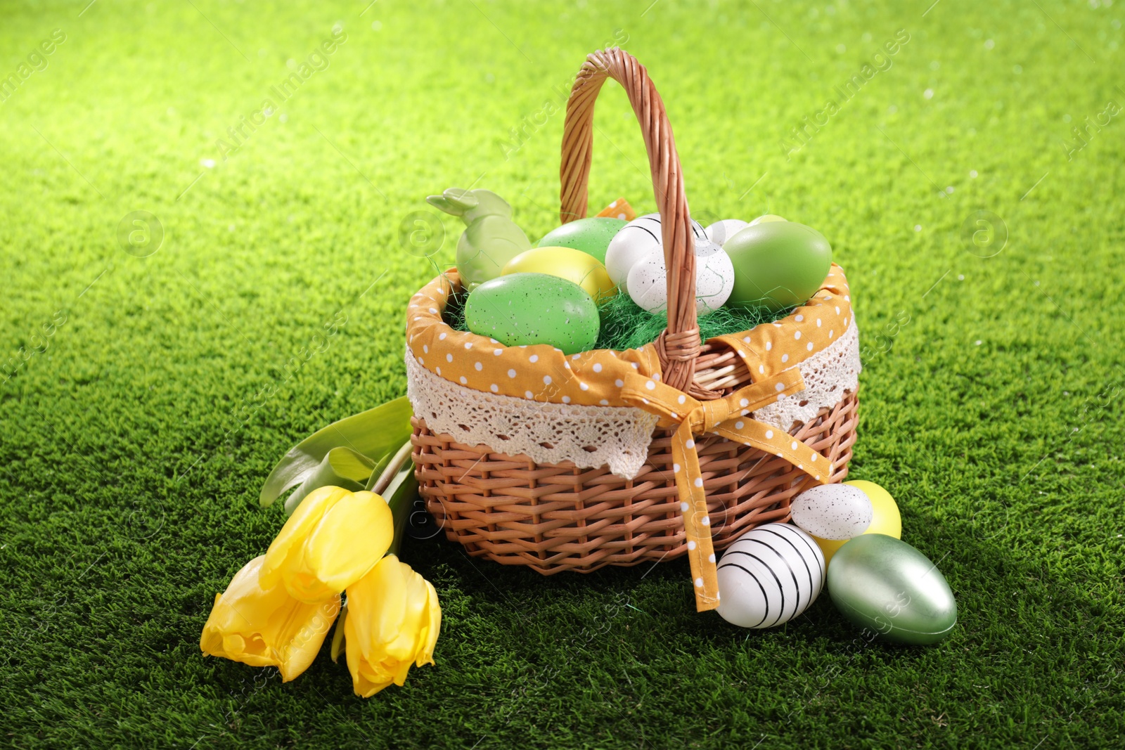 Photo of Easter basket with beautifully painted eggs, tulips and figure of rabbit on green grass