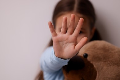 Photo of Child abuse. Little girl with teddy bear doing stop gesture on light grey background, selective focus. Space for text